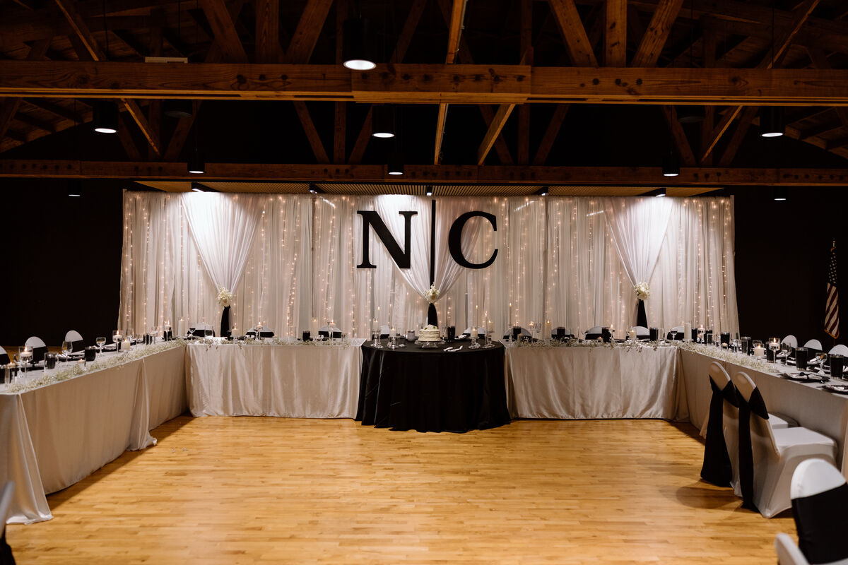 Reception head table with white drape backdrop and monogram letters lit with string lights inside the Eldora Ballroom